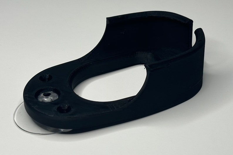 Puede incluir: Un soporte de pared de impresi&oacute;n 3D negro con una almohadilla adhesiva transparente en la parte inferior. El soporte tiene una forma curva y una abertura circular en el centro.