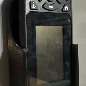 Puede incluir: Un soporte de pared de pl&aacute;stico negro para un buscador de peces de mano con una pantalla y botones. El soporte tiene un cable.