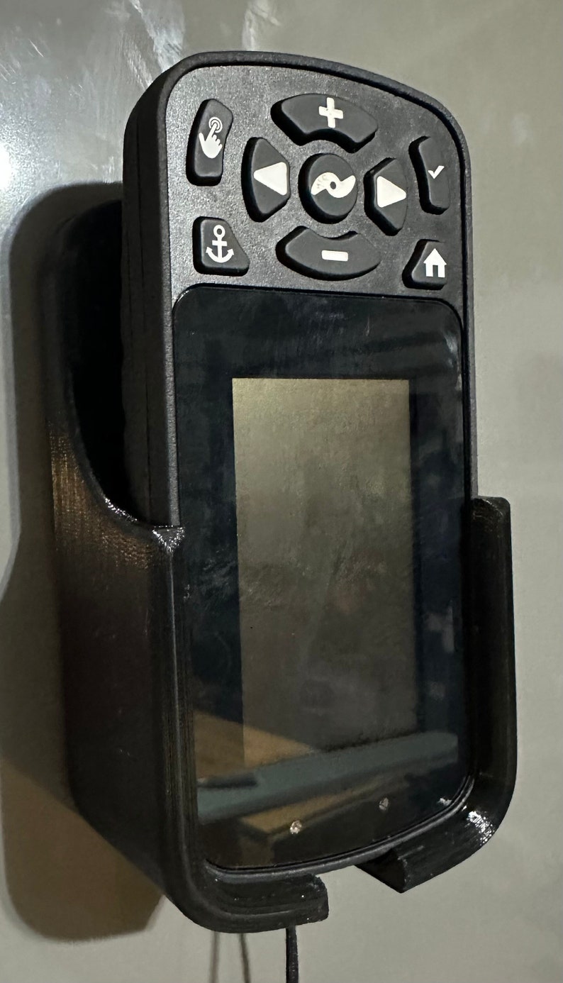 Puede incluir: Un soporte de pared de pl&aacute;stico negro para un buscador de peces de mano con una pantalla y botones. El soporte tiene un cable.