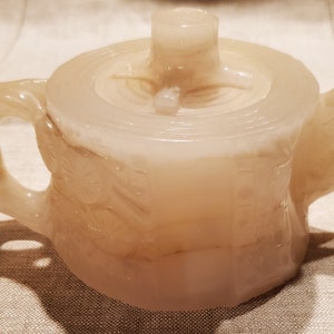 May include: A white ceramic teapot with a carved design. The teapot has a lid and a spout. The teapot is made in the shape of a tree trunk.