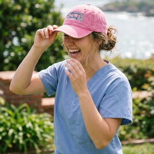 May include: A pink baseball cap featuring the embroidered text Caffeinated & Charting with a stethoscope graphic. The cap has a curved brim and a structured crown, with a washed-out fabric appearance and a small button on the top center.