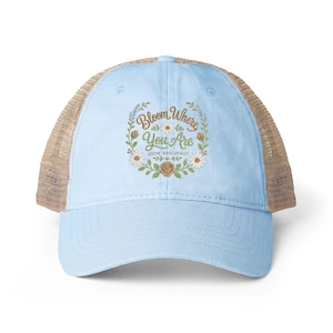 May include: A light blue baseball cap with a tan mesh back. The front features an embroidered floral wreath design in green, white, and gold, surrounding the text "Bloom Where You Are" and "GROW GRACEFULLY" in a cursive and serif font.