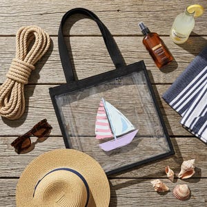 May include: A clear tote bag with a black frame and handles, featuring a sailboat illustration with pink and blue sails. The image also includes a straw hat, sunglasses, rope, a bottle of lemonade, and seashells, suggesting a beach theme.