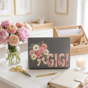 May include: A dark grey laptop skin featuring a floral bouquet of pink and white daisies tied with a gingham ribbon, alongside the name GIGI in large block letters filled with a pink floral pattern, displayed on a closed laptop surface.