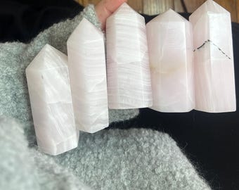 May include: Five pink and white crystal obelisks with a polished finish. The crystals are held in a hand, and the obelisks have a rectangular base and a pointed top. The crystals have a translucent appearance and are a light pink color.