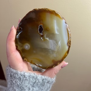 May include: A polished agate slice with a natural, organic shape. The stone features bands of brown, cream, and translucent white, with a dark, hollow cavity. The edges are a darker brown, and the overall appearance is smooth and glossy.