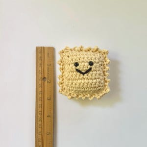 May include: A crocheted ravioli plush toy with a smiling face. The toy is yellow and has a black stitched smile and eyes.