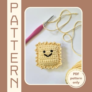 May include: A crocheted ravioli plush toy with a smiling face. The toy is yellow and has a ruffled edge. The image also shows a pink crochet hook and a ball of yarn.