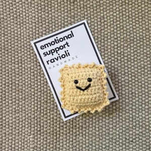 May include: Handmade crocheted ravioli with a smiling face. The ravioli is yellow and has a black crocheted smile and eyes. The ravioli is on a white card with the text "emotional support ravioli handmade".