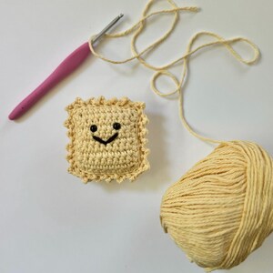 May include: A crocheted ravioli with a smiling face, a ball of yellow yarn, and a pink crochet hook.