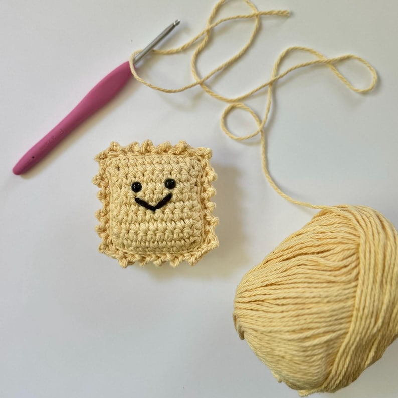 May include: A crocheted ravioli with a smiling face, a ball of yellow yarn, and a pink crochet hook.