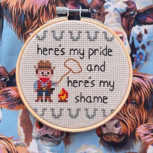 May include: A cross-stitch embroidery hoop with the text "here's my pride and here's my shame" and a small cowboy figure. The background is a fabric with a cow pattern.