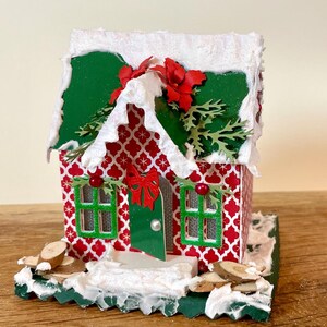 Könnte beinhalten: Ein Miniatur-Weihnachtshaus mit rot-weiß gemusterter Fassade, grünem Dach und schneebedeckten Akzenten. Das Haus hat grüne Fensterrahmen, eine rote Schleife und dekoratives Grün. Die Basis ist grün mit Kunstschnee und kleinen Holzstücken.