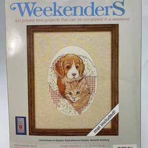 May include: A Weekenders counted cross stitch kit featuring a framed cross-stitch design of a dog and cat. The frame measures 8" x 10" (20 cm x 25 cm), and the finished size is 5" x 7" (13 cm x 18 cm). The kit includes a mat.