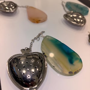 May include: A collection of tea infusers with gemstone accents. The infusers have a heart-shaped and shell-shaped design, connected to a chain and a teardrop-shaped gemstone. The gemstones are in shades of pink, blue, and yellow.