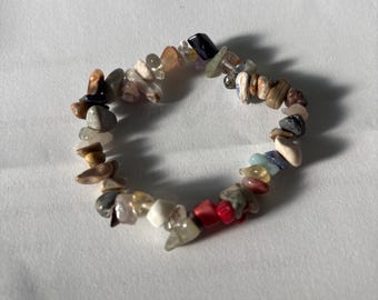 Puede incluir: Un brazalete colorido hecho de pequeñas piedras de forma irregular. Las piedras son de varios colores, incluyendo rojo, blanco, negro, marrón y azul. El brazalete está ensartado en una banda elástica.