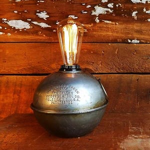 May include: A vintage-style lamp with a spherical metal base and a clear Edison bulb. The base has embossed text and a weathered, industrial aesthetic. The lamp sits on a wooden surface, creating a rustic ambiance.