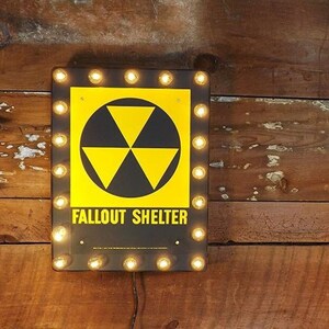 May include: A black and yellow fallout shelter sign with a glowing light bulb border. The sign has a black and yellow radiation symbol and the text "Fallout Shelter".