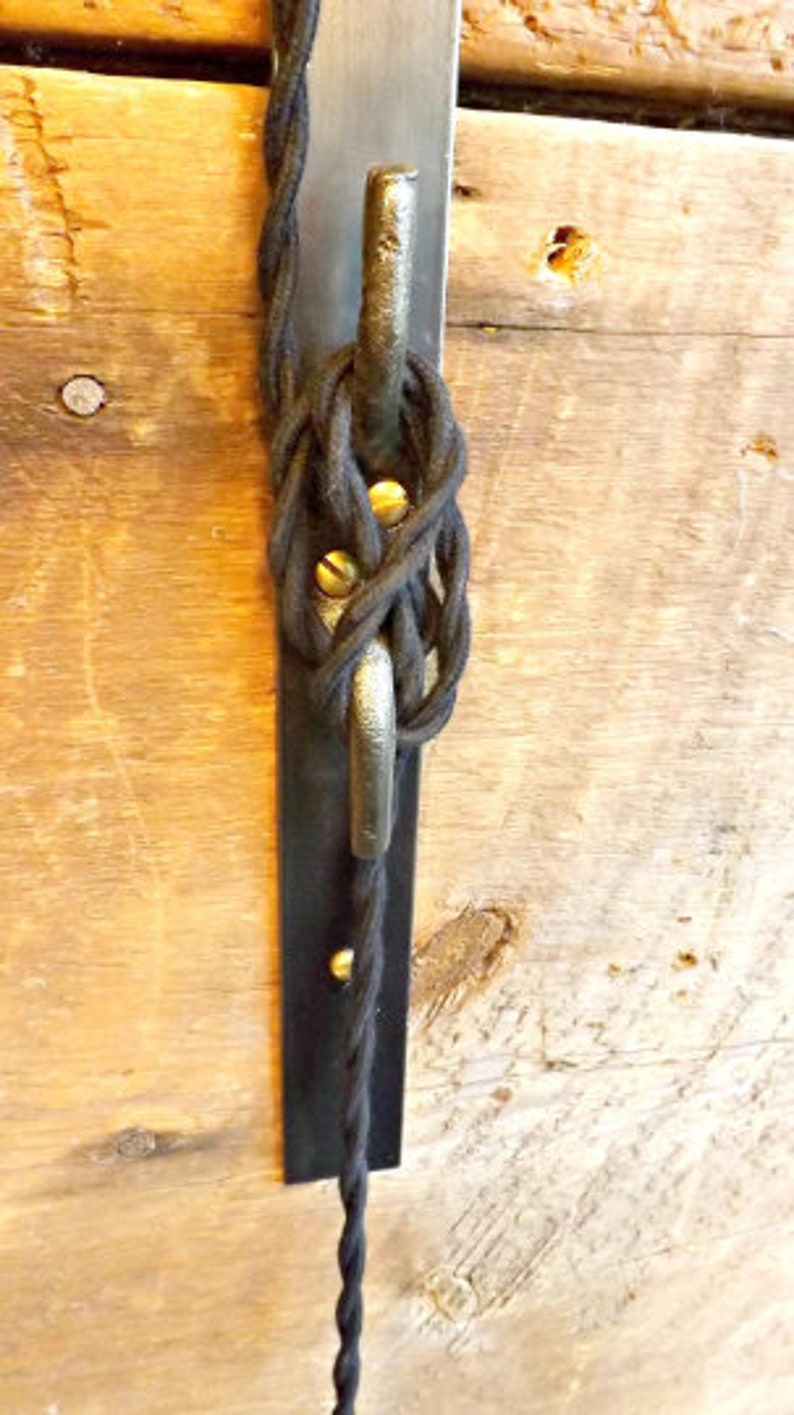 May include: Black electrical cord wrapped around a metal hook attached to a black metal plate. The plate is mounted to a wooden surface.