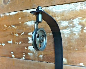 May include: A black metal pulley mounted on a curved bracket. The pulley has a round, black wheel with a small hole in the center.