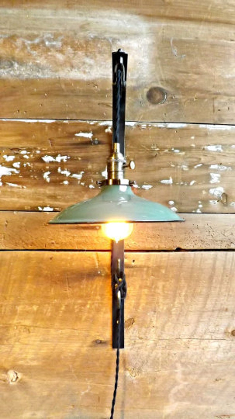 May include: A green metal lampshade with a black cord and a metal bracket attached to a wooden wall. The lampshade is hanging from the bracket and has a light bulb inside.