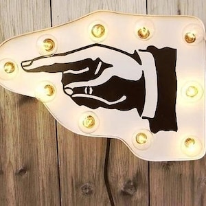 May include: White, arrow-shaped light-up sign featuring a black and white hand pointing to the right. The sign is outlined with warm, yellow light bulbs. The sign is mounted on a wooden background.