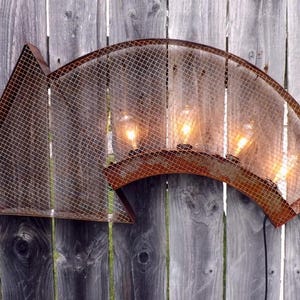 May include: A rustic, arrow-shaped wall light fixture. The metal frame has a distressed, rusty finish, with a wire mesh covering the light bulbs. The fixture is mounted on a weathered wooden fence.