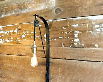 May include: A black metal wall-mounted light fixture with a pulley system and a hanging light bulb. The fixture is mounted on a wooden wall.