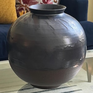 Könnte beinhalten: Eine weiße Keramik-Tasse mit schwarzem Henkel und dem Text "But First, Coffee" in fetter, schwarzer Schrift. Die Tasse steht vor einem verschwommenen Hintergrund, der eine gemütliche Umgebung suggeriert.