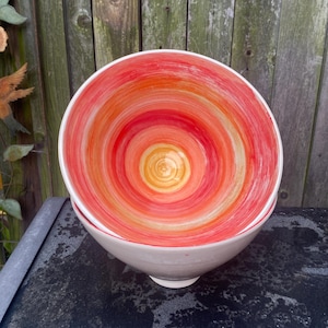 Puede incluir: Dos cuencos de cerámica blanca apilados, con un diseño interior vibrante. El interior de los cuencos presenta un patrón en espiral de tonos naranja, rojo y amarillo. Los cuencos están colocados contra una valla de madera desgastada.