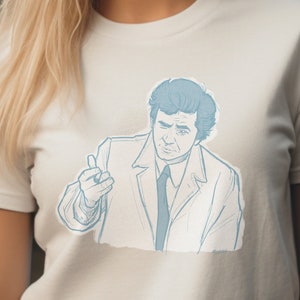 May include: A light beige t-shirt featuring a light blue sketch of a man in a suit, pointing with his finger. The illustration has a vintage, hand-drawn style. The t-shirt is made of a soft, comfortable material.