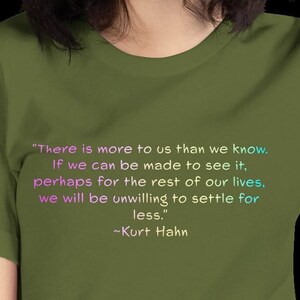 May include: A green t-shirt with a quote printed on the front in pink, blue, and white. The quote reads: "There is more to us than we know. If we can be made to see it, perhaps for the rest of our lives, we will be unwilling to settle for less." ~Kurt Hahn