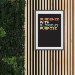 May include: A framed print with a black background and white text that reads "BURDENED WITH GLORIOUS PURPOSE". The text is in a retro font style with different colored letters.