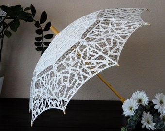 Puede incluir: Un parasol de encaje blanco con un mango y marco de madera. El intrincado diseño de encaje del parasol es visible, y está parcialmente abierto. El fondo incluye flores blancas y vegetación, lo que sugiere un tema decorativo o de boda.