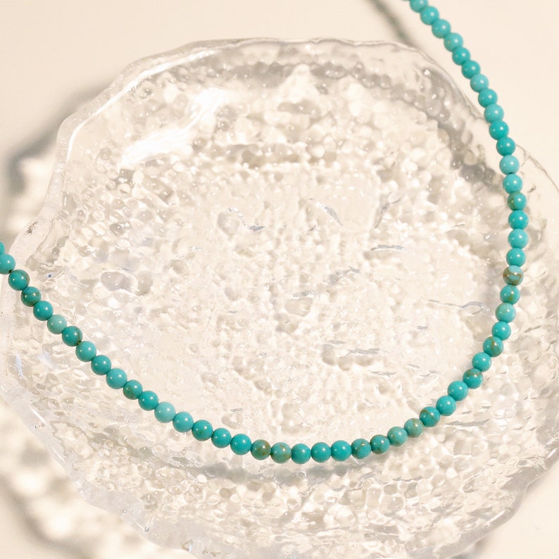 Puede incluir: Un collar de cuentas azul turquesa sobre una superficie de vidrio transparente.