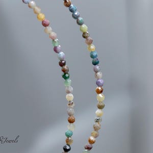 May include: A close-up of a necklace with small, colorful beads. The beads are in shades of pink, yellow, green, blue, and brown. The necklace is hanging against a blurred gray background. The brand name "Endi Jewels" is in the bottom left corner.