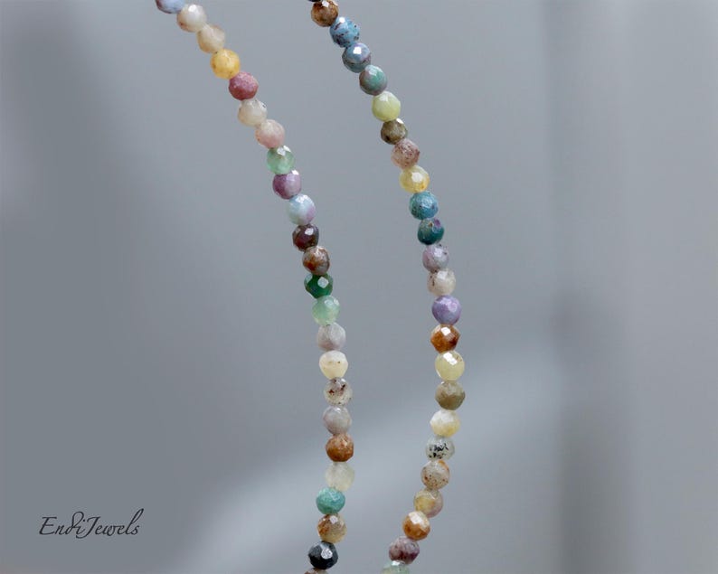 May include: A close-up of a necklace with small, colorful beads. The beads are in shades of pink, yellow, green, blue, and brown. The necklace is hanging against a blurred gray background. The brand name "Endi Jewels" is in the bottom left corner.