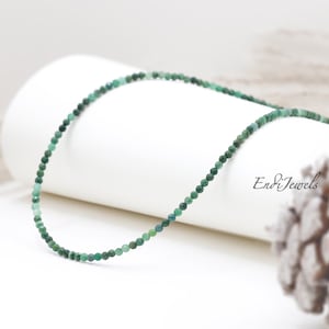 May include: A delicate green gemstone necklace made with small, round beads. The necklace is on a white surface with a pine cone in the background.