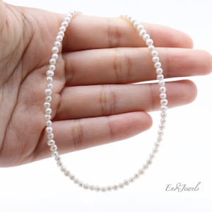 Może przedstawiać: Delikatny biały naszyjnik z pereł o prostym wzorze. Naszyjnik wykonany jest z małych, okrągłych pereł, które są nawleczone na cienki łańcuszek.