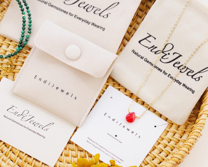 May include: An assortment of jewelry pieces, including a gold chain necklace with a red gemstone pendant and a green beaded necklace, are displayed on a woven tray. The jewelry is packaged in small, off-white pouches and cards with the brand name "Endi Jewels".