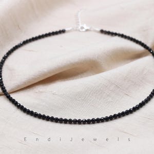 K&ouml;nnte beinhalten: Eine zarte Halskette mit kleinen, facettierten schwarzen Perlen, die kreisf&ouml;rmig angeordnet sind. Das Schmuckst&uuml;ck verf&uuml;gt &uuml;ber einen silberfarbenen Metallverschluss und eine verstellbare Verl&auml;ngerungskette. Der Text Endi Jewels steht unten mittig.
