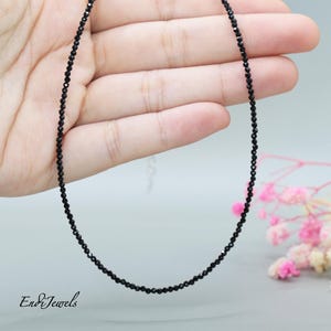 Puede incluir: Un delicado collar negro hecho con pequeñas cuentas de espinela negra facetadas. El collar se muestra en una mano, con las cuentas formando una cadena simple y elegante.