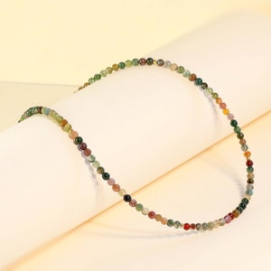 May include: A multi-colored beaded necklace with small, round beads in shades of green, brown, red, and gold. The necklace is made of natural stone beads and is strung on a thin gold chain.