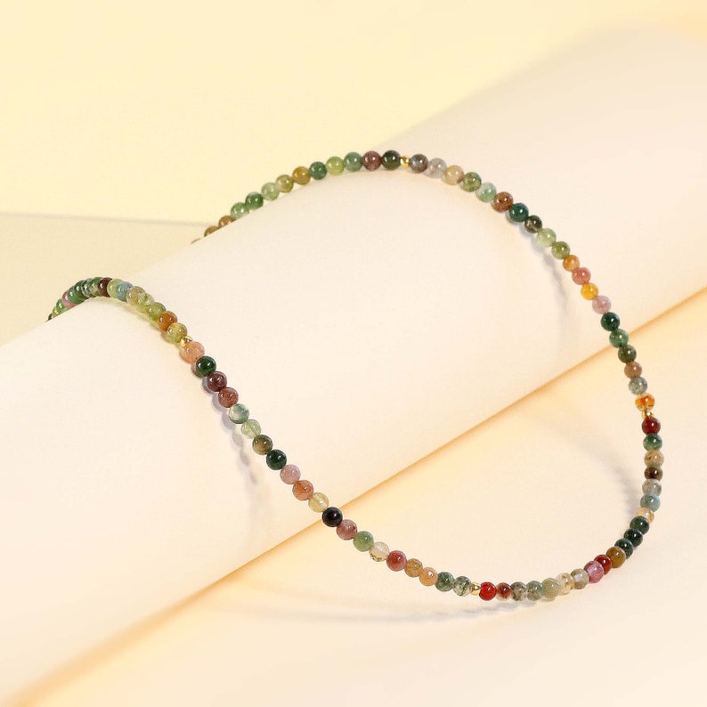 May include: A multi-colored beaded necklace with small, round beads in shades of green, brown, red, and gold. The necklace is made of natural stone beads and is strung on a thin gold chain.