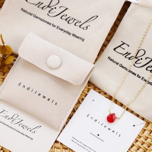 May include: A red gemstone necklace with a gold chain and a small red heart pendant. The necklace is displayed on a white velvet pouch with the text "Endi Jewels" and "Natural Gemstones for Everyday Wearing".