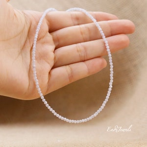May include: Delicate, light blue beaded necklace. The necklace is composed of small, round beads strung together, creating a subtle, elegant design. The necklace is shown against a neutral background, highlighting its delicate nature. The brand name "En&Jewels" is visible.