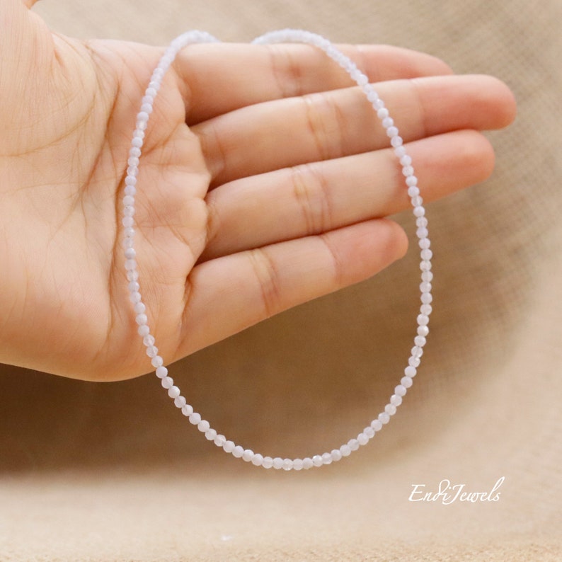 May include: Delicate, light blue beaded necklace. The necklace is composed of small, round beads strung together, creating a subtle, elegant design. The necklace is shown against a neutral background, highlighting its delicate nature. The brand name "En&Jewels" is visible.