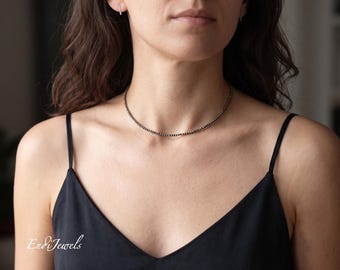 May include: A fine beaded necklace featuring small, dark beads. The necklace is worn around the neck, complemented by a black camisole. The image also shows small hoop earrings. The jewelry is from Endi Jewels.