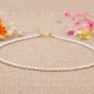 May include: A delicate white pearl necklace with a gold clasp. The necklace is on a brown textured surface.