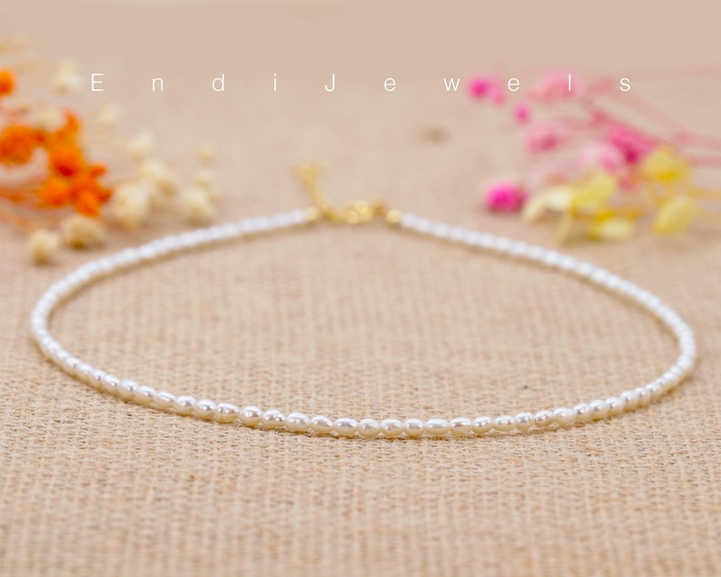 May include: A delicate white pearl necklace with a gold clasp. The necklace is on a brown textured surface.
